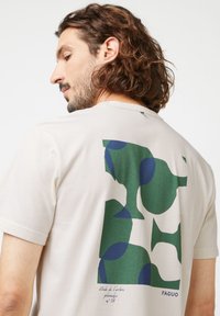 Beige T-Shirt mit einem großen abstrakten Grafikdesign in Grün und Blau auf dem Rücken. Aus weichem Material mit Rundhalsausschnitt und kurzen Ärmeln.