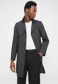 Ciemnoszary płaszcz trencz z podwójnym rzędem guzików, ostrym kołnierzem, długimi rękawami, para czarnych spodni, noszony na jasnej koszuli.