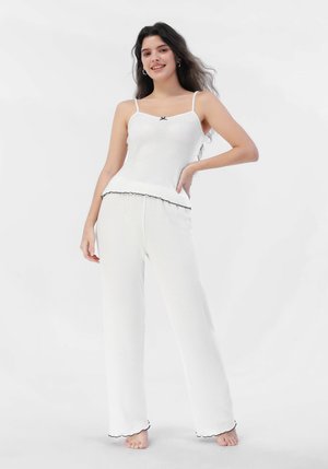 Camiseta de tirantes finos de canalé blanca con un detalle de lazo negro, combinada con pantalones anchos blancos con dobladillos en forma de ondas. Textura suave.