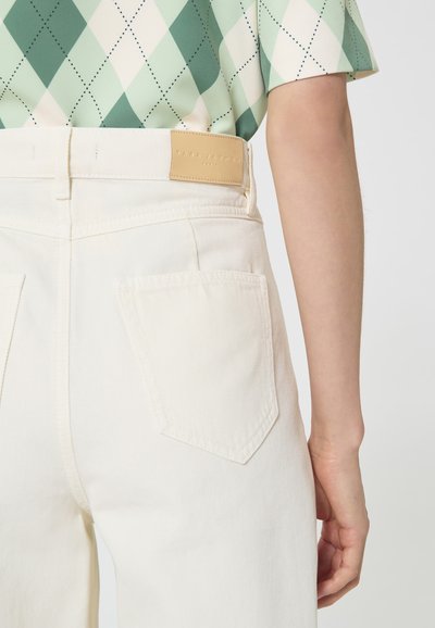 Personne portant un jean taille haute couleur blanc cassé avec des poches arrière et une chemise à motif losanges verts et blancs rentrée à l'intérieur.