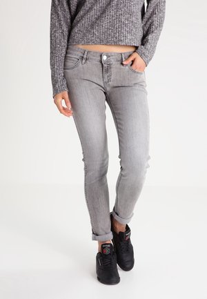 Person, die graue Skinny-Jeans mit umgekrempelten Bündchen, einen grauen Strickpullover und schwarze Reebok-Sneakers trägt, steht mit einer Hand in der Tasche.
