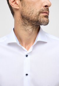 Weißes Hemd mit einem kontrastierenden gemusterten Innenkragen, zwei schwarzen Knöpfen und einem klaren Design mit glatter Textur.