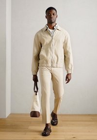 Μπεζ φούτερ με φερμουάρ, κρεμ πουκάμισο, κρεμ παντελόνι και καφέ μοκασίνια. Κρατάει μια λευκή τσάντα με καφέ λεπτομέρειες. Ξύλινο δάπεδο και ουδέτερο φόντο.