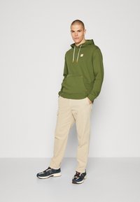 Olivengrønn hettegenser med hvite snorer, frontlomme i kengurustil, paret med beige cargo-bukser og marineblå joggesko.
