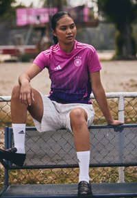 Maillot de sport en dégradé rose et violet avec un col rond et trois rayures, associé à un short blanc rayé et des chaussures noires.