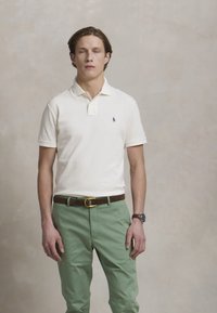 Ung man med slutna ögon, iförd vit polo, gröna byxor, brunt bälte och klocka på handleden, stående mot en enfärgad beige bakgrund.