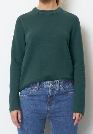 Kobieta ubrana w teksturowaną ciemnozieloną bluzkę z długim rękawem, lekko wpuszczoną do wysokich niebieskich dżinsów na jednolitym tle.