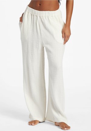 Mujer con pantalones blancos sueltos, texturizados y de pierna ancha con cintura elástica y manos en los bolsillos, descalza sobre un fondo blanco.