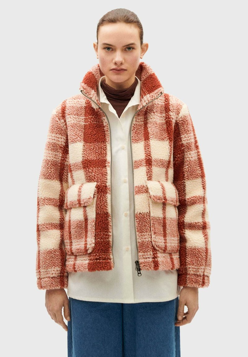 Red and cream checkered fleece jacket with a zip front, high collar, two front pockets, and soft texture. Worn over a white shirt.