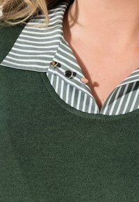 Maglione verde a maglia con un colletto a righe bianche e verdi. Due bottoni decorativi sul colletto, realizzati in tessuto testurizzato.