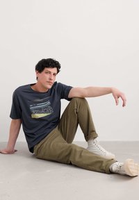 Camiseta marina con un diseño gráfico, pantalones verde oliva y zapatillas blancas. La camiseta presenta arte lineal abstracto y sutiles acentos de color.