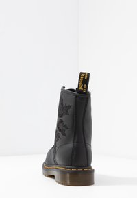 Bottine en cuir noir ornée de motifs floraux brodés, équipée d'une boucle jaune à l'arrière et d'une épaisse semelle en caoutchouc transparente.