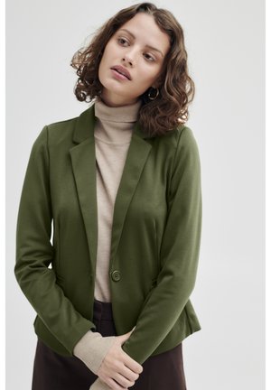 Tmavě zelený blazer s úzkým střihem, s jedním knoflíkem a klopovými límci. Nosený přes béžový rolák, který má hladkou tkaninu a přizpůsobený design.