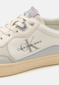 Calvin Klein tenisice s krem kožnim gornjim dijelom s sivo-zlatnim detaljima, teksturiranom završnom obradom, veznim dizajnom i bež gumiranim potplatom.