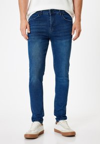 Jeans de mezclilla azul con corte ajustado, cinco bolsillos y un lavado desgatado. Presenta un cierre de botón y trabillas para cinturón. Combinados con zapatillas blancas.