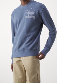 Modrý crewneck svetr vyrobený z bavlny, se vyšitým logem Adidas a designem tří pruhů na hrudi. Měkká textura.