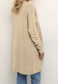 Long gilet beige en tricot avec un motif en chevrons porté sur un jean bleu, vu de dos et de côté.