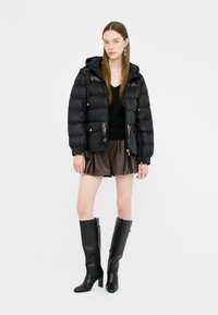 Giovane donna che indossa un giubbotto a vento nero, un maglione nero, una gonna in pelle marrone e stivali neri a ginocchio, in piedi contro uno sfondo bianco.