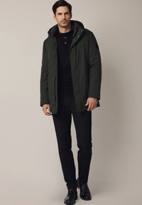 Mężczyzna w ciemnozielonej kurtce z kapturem, na czarnym swetrze, czarnych spodniach i czarnych botkach na kostkę, na jasnobeżowym tle.