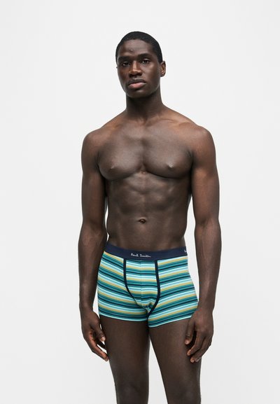 Homme musclé debout sur fond blanc, portant un boxer rayé bleu et vert avec une ceinture Paul Smith.