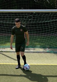 Zaļā futbola forma ar logo, melnās zeķes un futbols. Spēlētājs stāv pie rakstainā bumbas priekšā vārtiem ar zaļu tīklu.