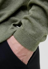 Zaļš adīts džemperis, kura piedurkne balstās uz melna kabatas, izceļot tekstūras audumu un ribu apakšmalu, ar smalku spīdumu.