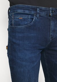 Mörkblå denimjeans med slim-fit-design, femficksstil och en synlig logotagg med orange detaljer på baksidan.