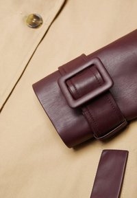 Tessuto beige con una cinghia in pelle bordeaux scuro dotata di fibbia rettangolare. La texture è liscia con dettagli di cuciture a contrasto.