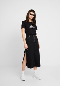 Jupe midi en denim noir avec fentes latérales et coutures contrastées, associée à un t-shirt noir avec logo blanc et manches courtes. Baskets blanches.