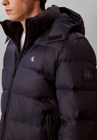 Homme portant une doudoune noire Calvin Klein avec fermeture éclair à l'avant et capuche sur un fond gris uni.