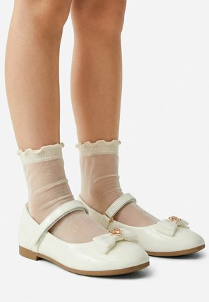 Nogi dziecka w przezroczystych kremowych skarpetkach z falbankami do kostek oraz białych lakierowanych butach typu Mary Jane z kokardkami i złotymi serduszkami.