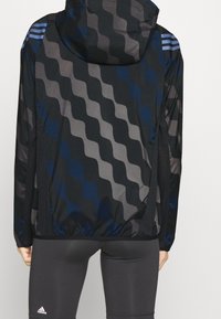 Giacca a vento con motivo a onde blu e grigio, realizzata in tessuto leggero, con cappuccio, orlo elastico e inserti in rete sui lati.