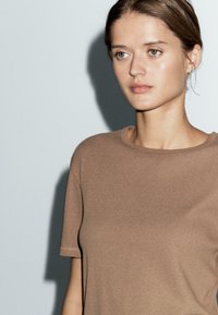Donna con pelle chiara e occhi verdi, che indossa una semplice maglietta marrone a maniche corte, mentre guarda leggermente di lato su uno sfondo chiaro e uniforme.