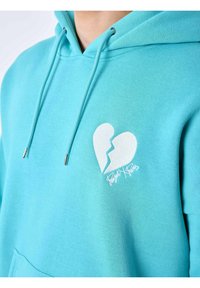 Sudadera color turquesa hecha de tela suave, con un gráfico de un corazón roto en blanco y el texto "Project Paris" bordado en el delantero. Incluye cordones.
