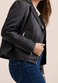 Veste de moto en cuir noir avec poches zippées, silhouette ajustée et détails en métal argenté. Portée sur un haut blanc et un jean bleu.