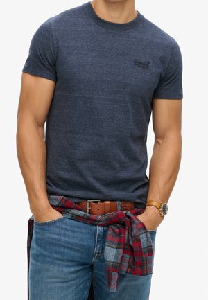 Mężczyzna w dopasowanym ciemnoniebieskim t-shircie z czerwoną i niebieską koszulą w kratę zawiązaną wokół talii, niebieskich dżinsach, brązowym pasku i złotym zegarku.