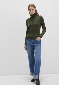 Dunkelgrüner Strickpullover mit Rollkragen, kombiniert mit hellblauen weit geschnittenen Jeans mit umgeschlagenem Saum, getragen mit schwarzen Absatzstiefeln.