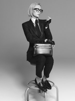 Black leather handbag with structured design, paired with a form-fitting suit, white shirt, and black tie. Gloves and chunky glasses complete the look.