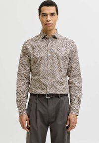 Homme portant une chemise beige à fleurs cintrée à manches longues, rentrée dans un pantalon gris plissé avec une ceinture noire, debout devant un fond uni.
