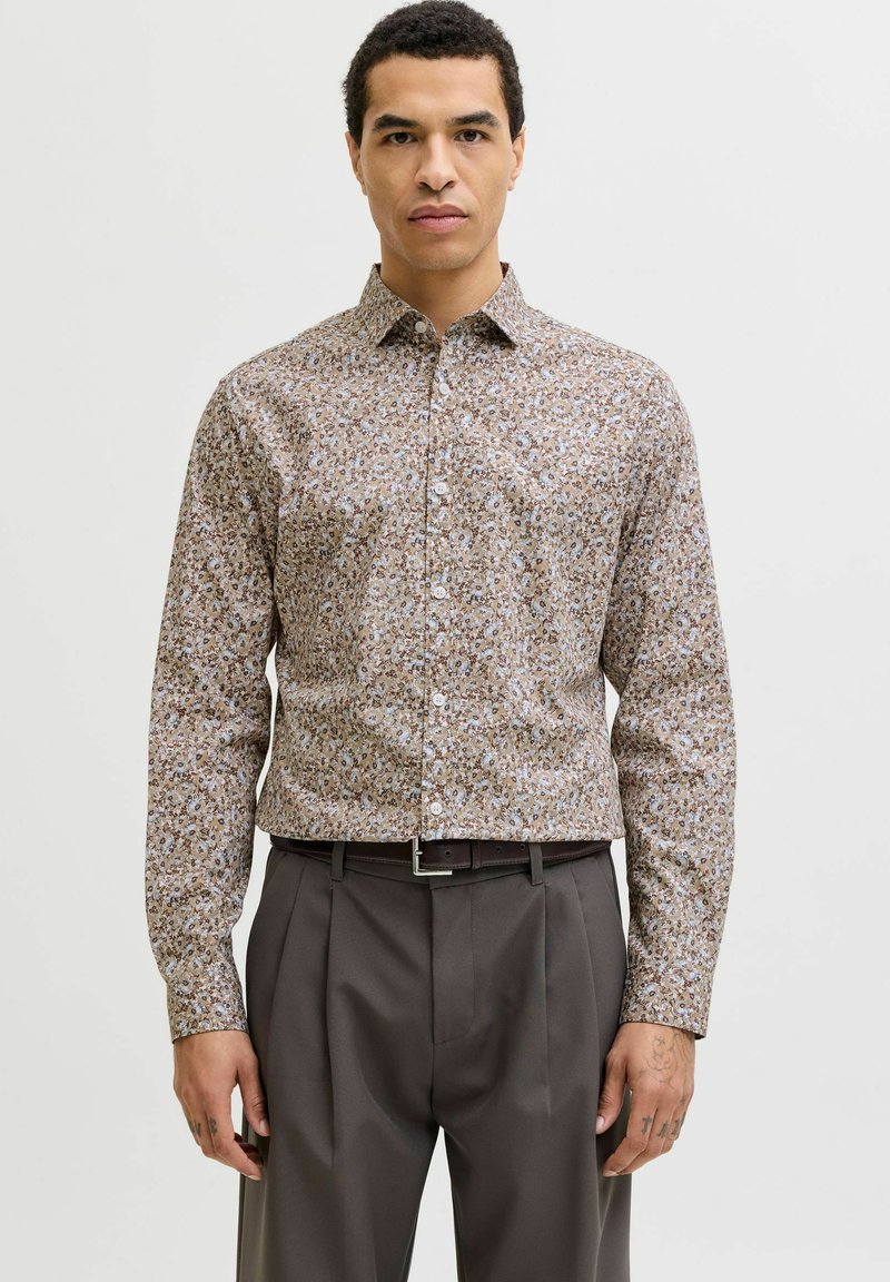 Homme portant une chemise beige à fleurs cintrée à manches longues, rentrée dans un pantalon gris plissé avec une ceinture noire, debout devant un fond uni.