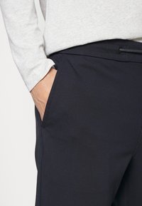 Marineblauwe broek heeft een gladde textuur, zijzakken en een verstelbare tailleband, gedragen met een lichtgrijze langslavenshirt.