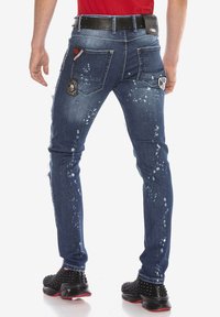 Jean en denim avec un lavage foncé, accents usés, motif éclaboussé, poches arrière doubles et détail de patch brodé. Associé à des chaussures noires à studs.