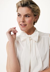 Blusa crema con colletto a fiocco, dettagli plissettati sulle spalle e texture morbida. Modella dai capelli corti tocca il mento.