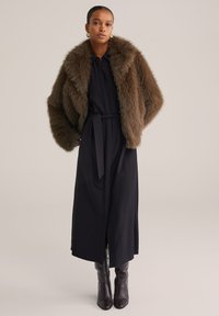 Vrouw in een bruine faux fur jas over een lange zwarte jurk met riem en donkerbruine kniehoge laarzen op een lichte achtergrond.