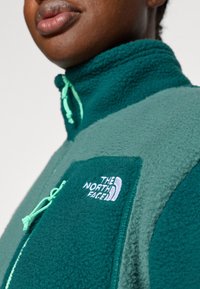 Teal fleecejakke med høy krage, glidelås foran og lomme. Med kontrasterende grønne og blå paneler, merket med logo i hvitt.