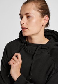 Svart huvtröja med dragsko, med en mjuk textur och raglanärmar. Modellen håller i dragskon nära kragen.