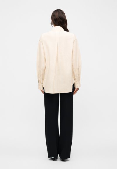 Γυναίκα, όρθια με την πλάτη γυρισμένη, φοράει μια μπεζ μπλούζα με μακριά μανίκια και διακριτικές ρίγες και φαρδιά μαύρη παντελόνα.