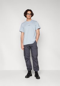 T-shirt en coton bleu clair avec un col rond, associé à un pantalon cargo gris foncé comportant plusieurs poches et des bottes noires à lacets.