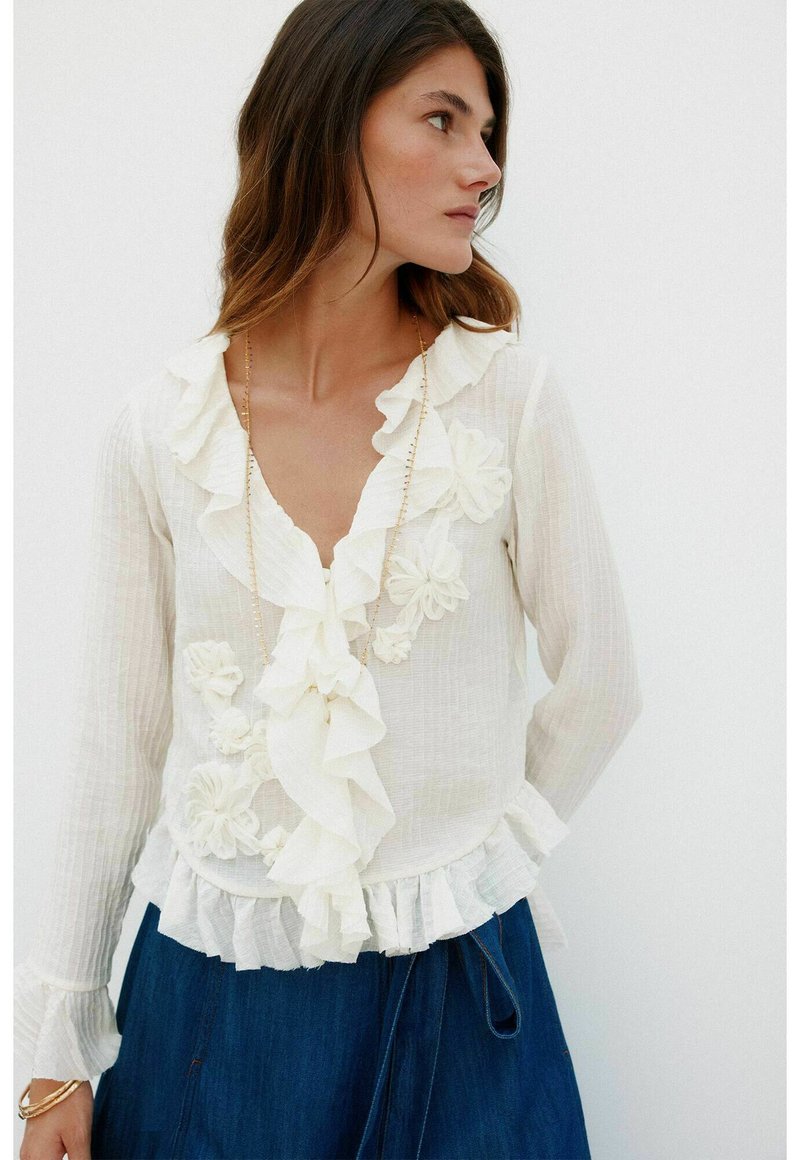 Weiße Bluse mit Rüschkragen und floralen Akzenten, strukturiertem Stoff, langen Ärmeln und einem fransigen Saum, kombiniert mit einem blauen Jeansrock.