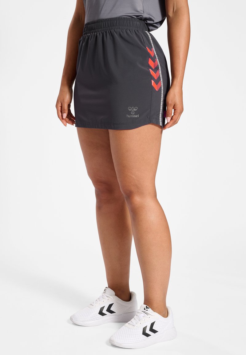 Sportieve rok in donkergrijs met een elastische tailleband, voorzien van rode chevron-accenten en reflecterende biezen. Gecombineerd met witte mesh sneakers.